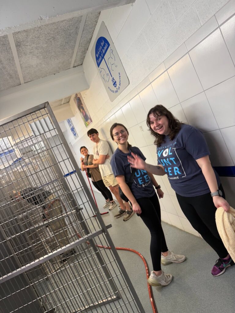 Group of volunteers standing in a clean, tiled hallway at an animal shelter, smiling and waving near kennel cages.