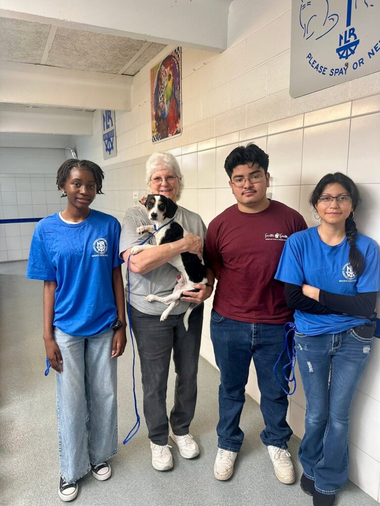 Four people stand indoors with a small tricolor dog at an animal shelter.