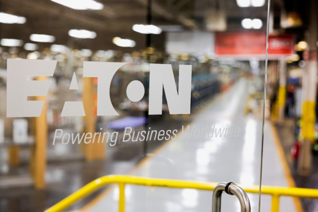 Glass door with the Eaton logo and slogan “Powering Business Worldwide” etched in white. The background shows an industrial facility with bright lighting, yellow safety railings, and machinery along a polished walkway.